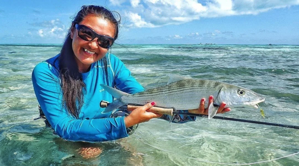 Smiling woman wearing Maui Jim Barrier Reef Sunglasses with Grey Mirror polarised lenses holding a fish and fly fishing rod in the ocean. | Best fly fishing sunglasses | Jumping Rise